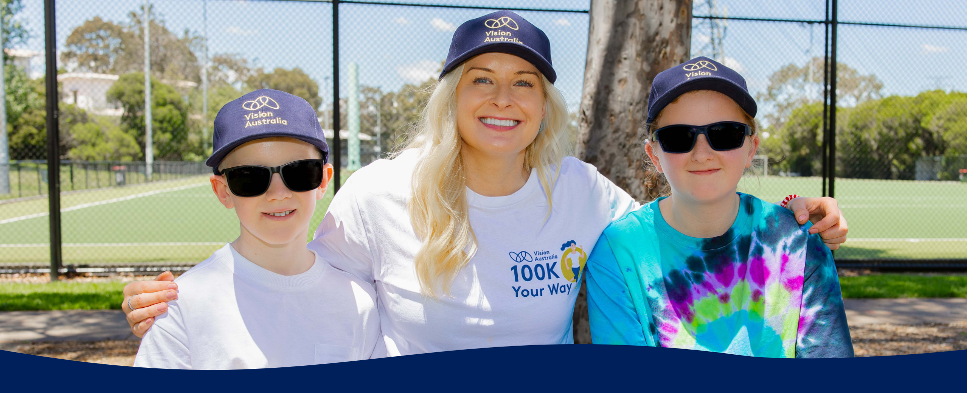 Woman stands between two children smiling wearing blue Vision Australia hats
