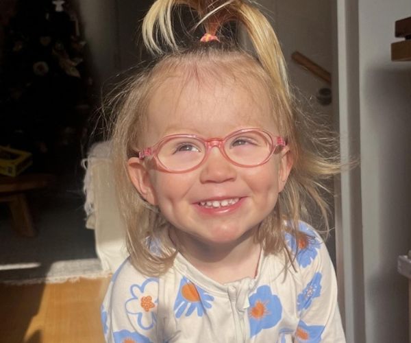 Sienna, a young girl wears a floral top, pink glasses, smiling