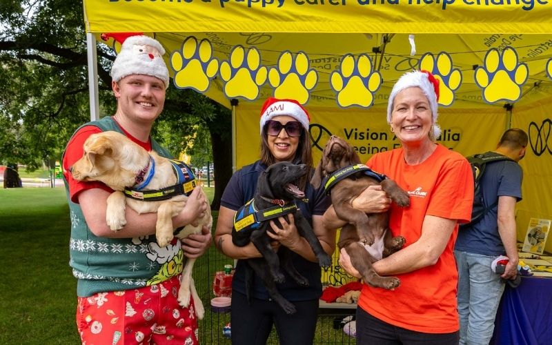 Three people wearing Santa hats stand in front of a Seeing Eye Dogs stand holding a Seeing Eye Dogs pup each smiling