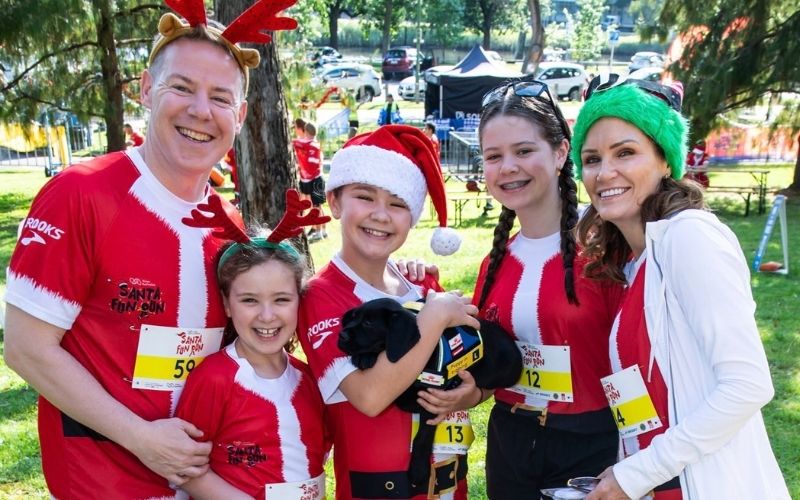 A family of five wearing Santa Fun Run tshirts, Christmas headwear stand together with a Seeing Eye Dogs puppy