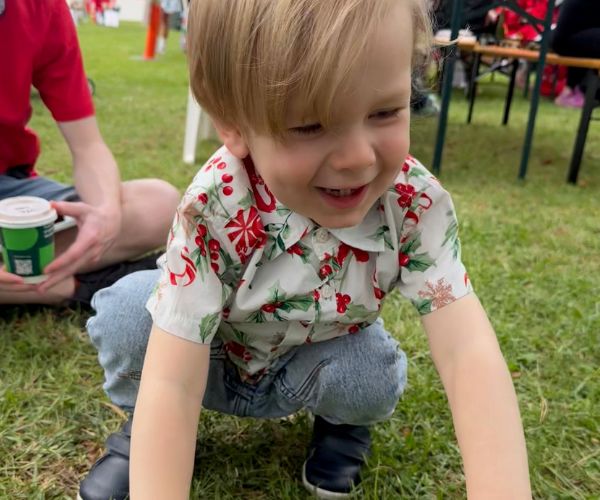 Young boy wearing a Christmas crouches down on the grass at Santa Fun Run event village