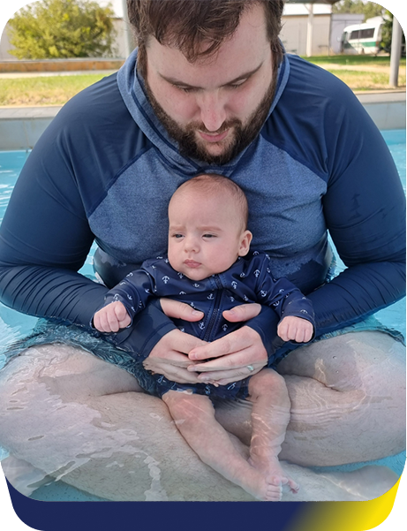 A man wearing a blue long sleeve top sits in a pool holding a baby also wearing blue