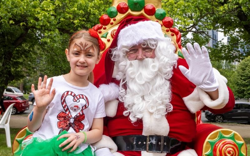 Girl wearing Christmas tshirt and her face painted sits with Santa on a throne waving