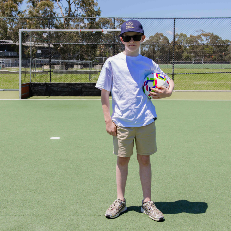 Vision Australia client Alfie holds a soccer ball and poses for a photo Infront of the goals.