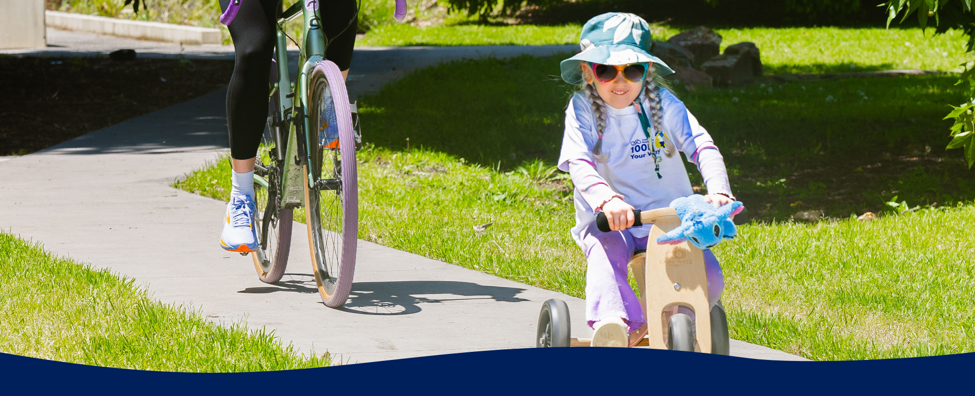 Girl wearing white 100K Your Way branded tshirt rides a trike on a path