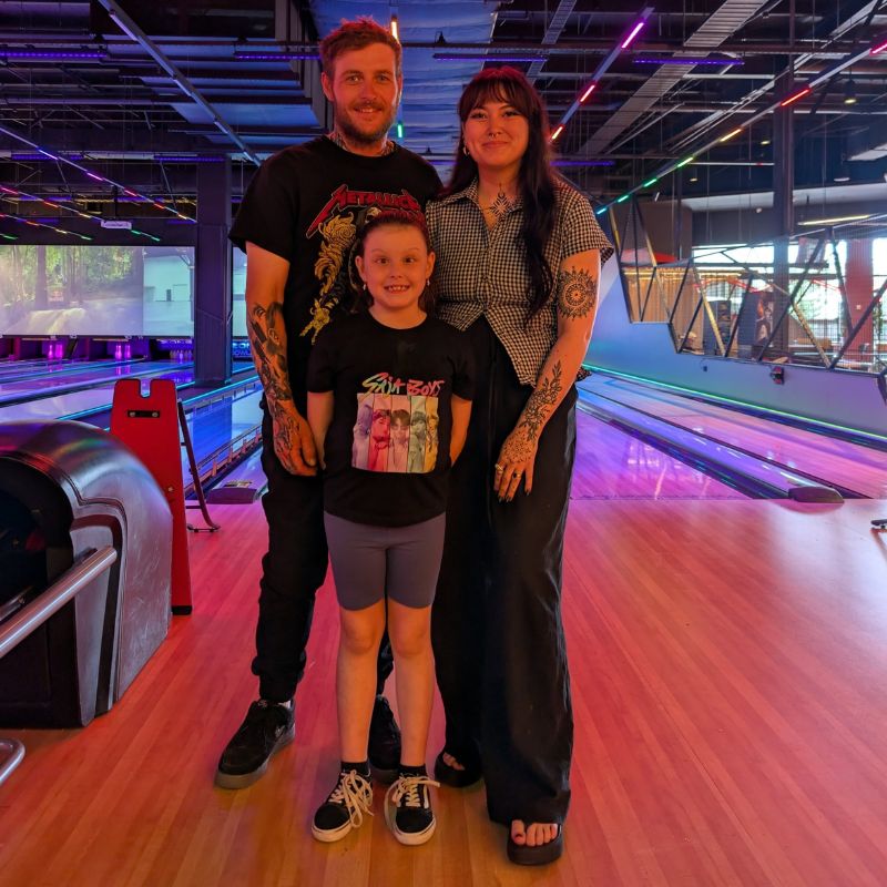Luna stands in front of her mum and dad in front of ten pin bowling lanes
