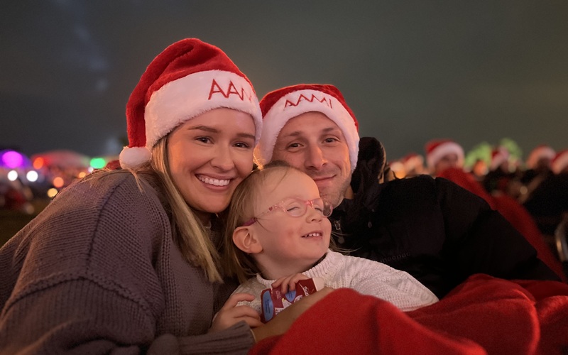 A woman and man wearing Santa hats hold a young girl wearing glasses in the Carols by Candlelight audience