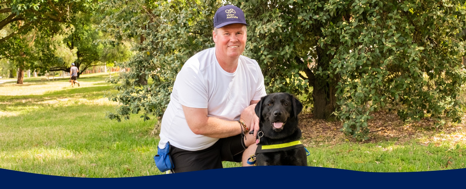 Man wearing white tshirt and blue Vision Australia cap kneels down next to black Seeing Eye Dog in a park smiling