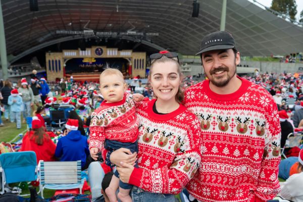 Two parents and a baby stand in the Carols audience wearing matching christmas jumpers