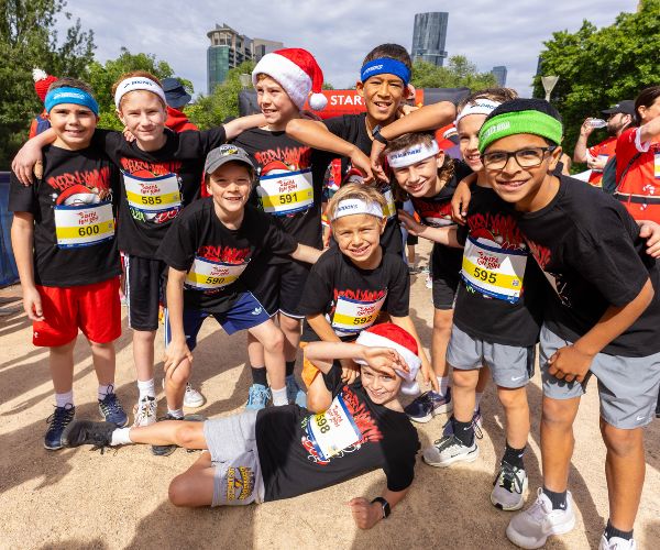 A group of 10 kids wearing Christmas tshirts and Santa Fun Run bibs huddle together and pose for the camera