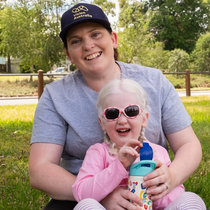 Jess in a cap holding her daughter Lina in her lap while sitting on the grass