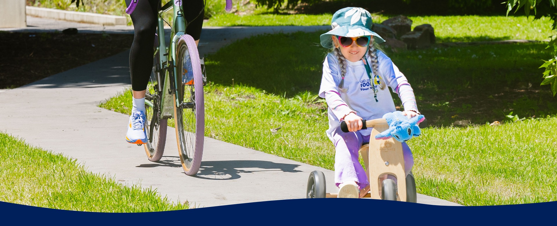 Girl wearing white 100K Your Way branded tshirt rides a trike on a path