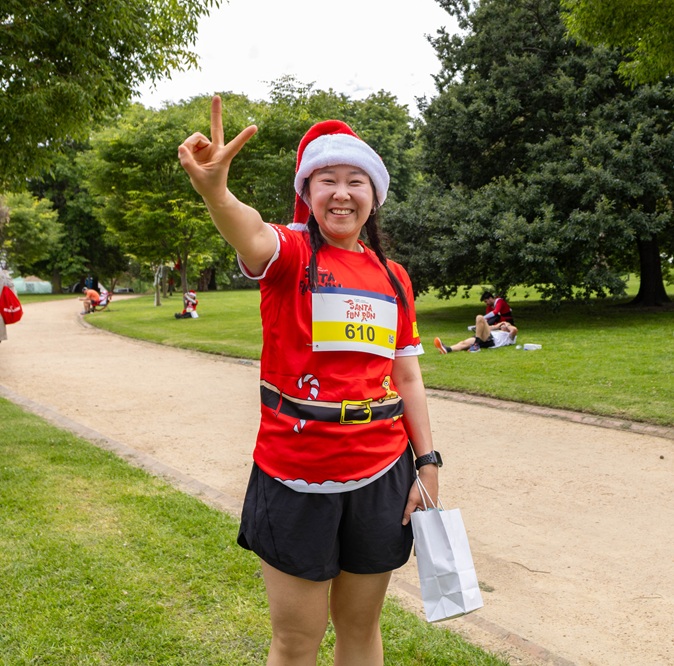 Lady wearing Santa Fun Run shirt and Santa Hat gives a peace sign beside the Santa Fun Run track