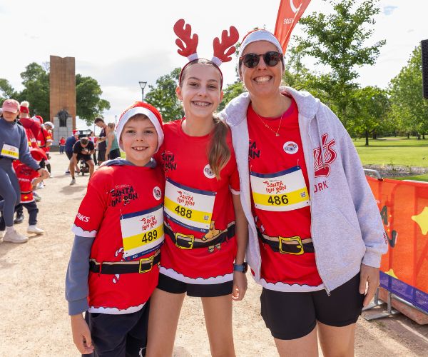 A woman and two kids wearing Santa Fun Run tshirts have their arms around eachother on the Santa Fun Run track smiling