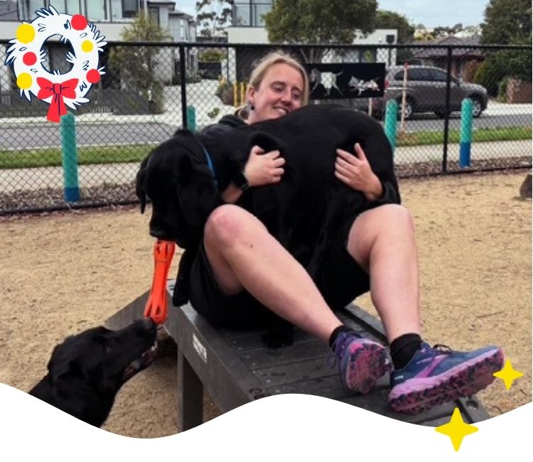 Woman sits on a ledge in a part with a black labrador in her lap and another on the ground next to her