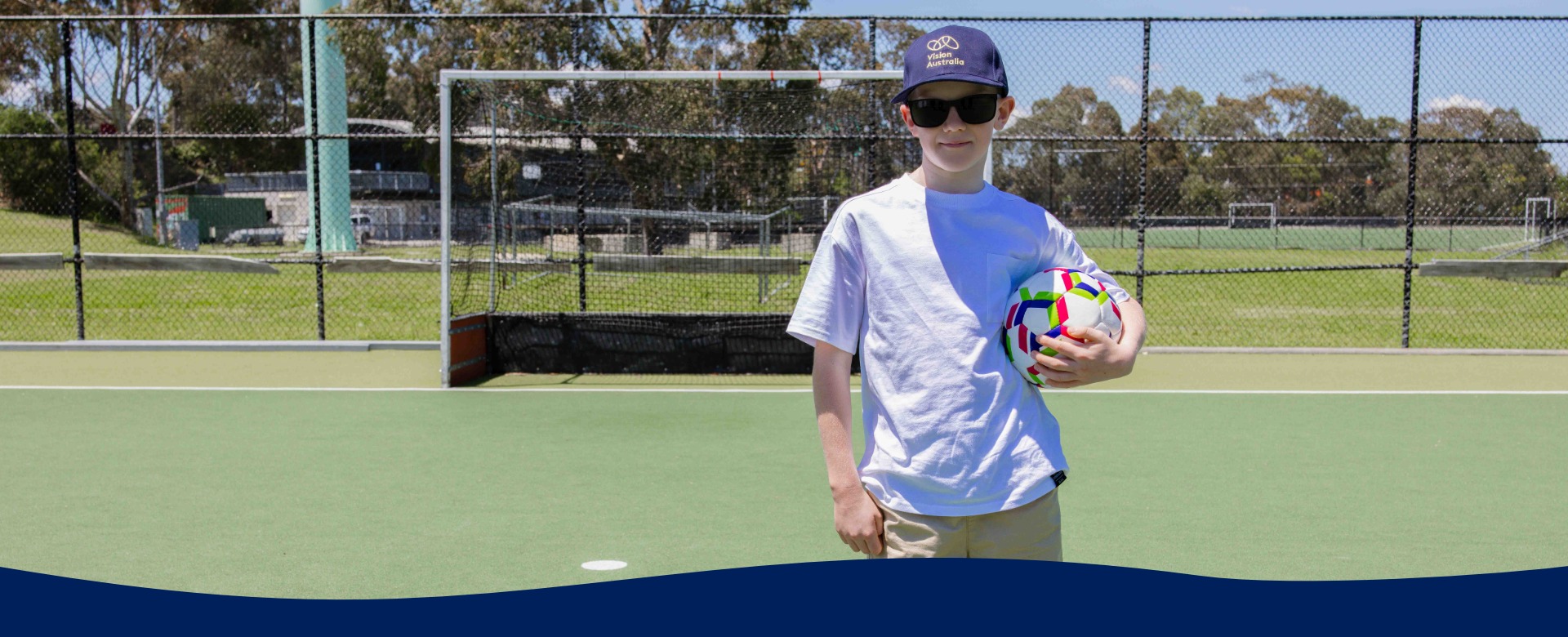 A boy wearing a white tshirt and a blue Vision Australia hat holds a soccer ball on a court