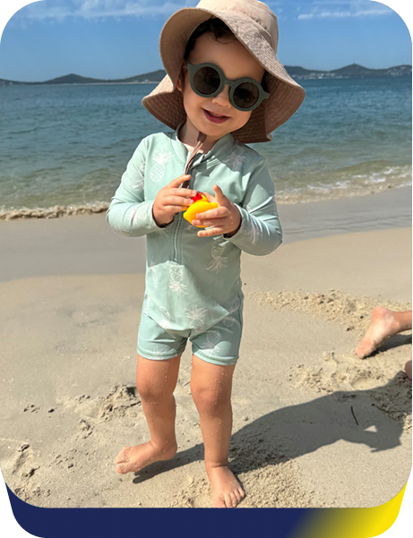 Young boy wearing a swim suit, glasses and a floppy hat stands on the beach smiling, holding a rubber ducky