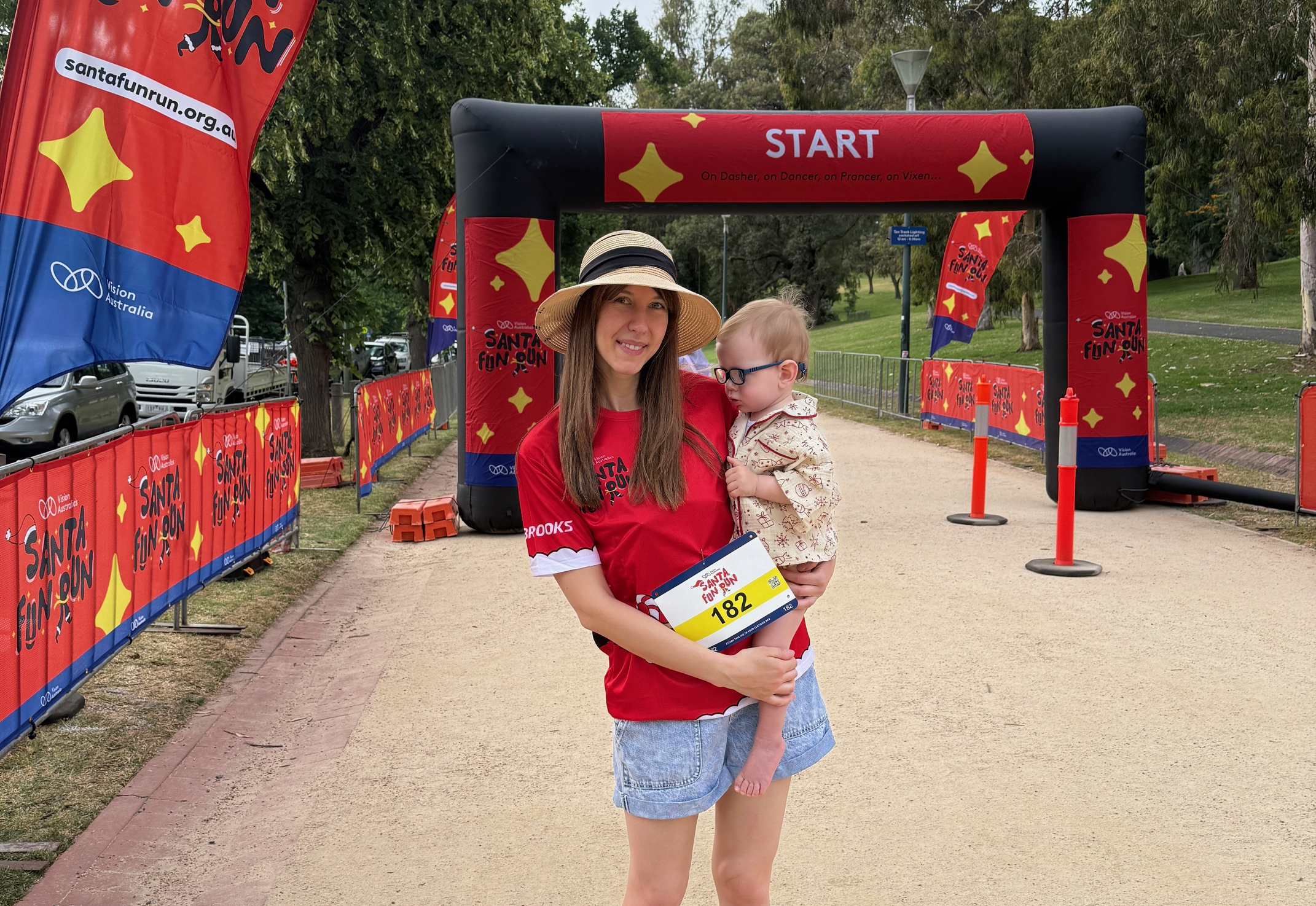 Woman wearing Santa Fun Run tshirt and straw hat holds young boy wearing glasses in front of the Santa Fun Run start line