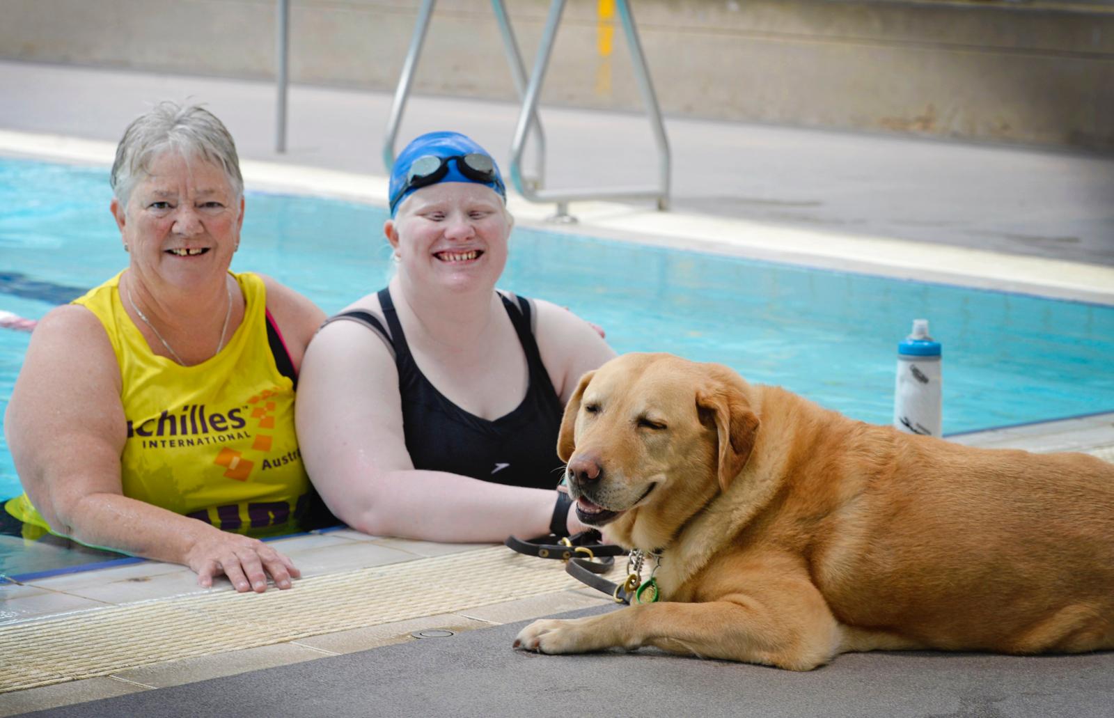 Two women in a pool with a dog lying near the edge